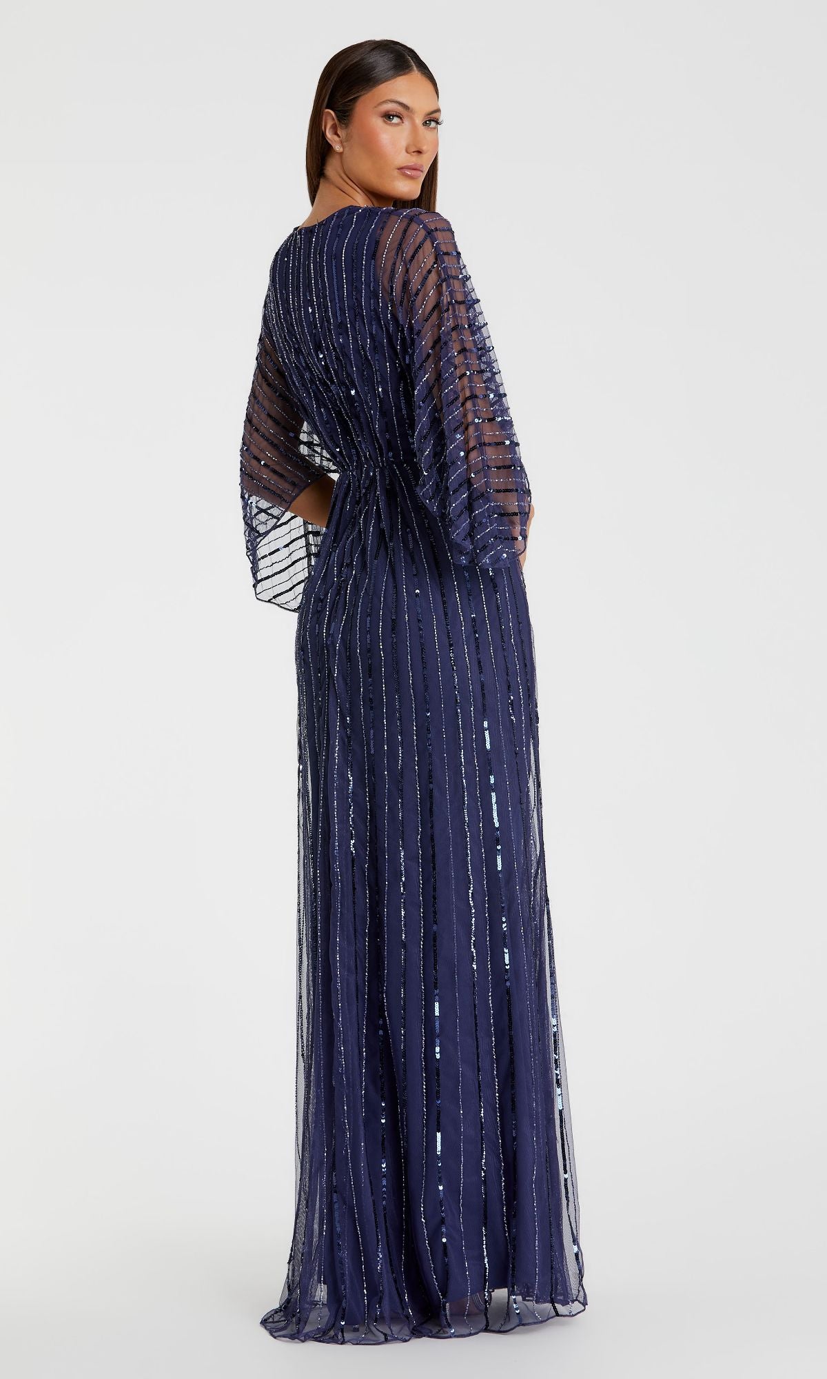 A woman in the Mac Duggal 93841 Long Sequin A-Line Formal Dress, featuring floor-length navy blue sheer sequined fabric and long sleeves, stands facing away while looking over her shoulder.